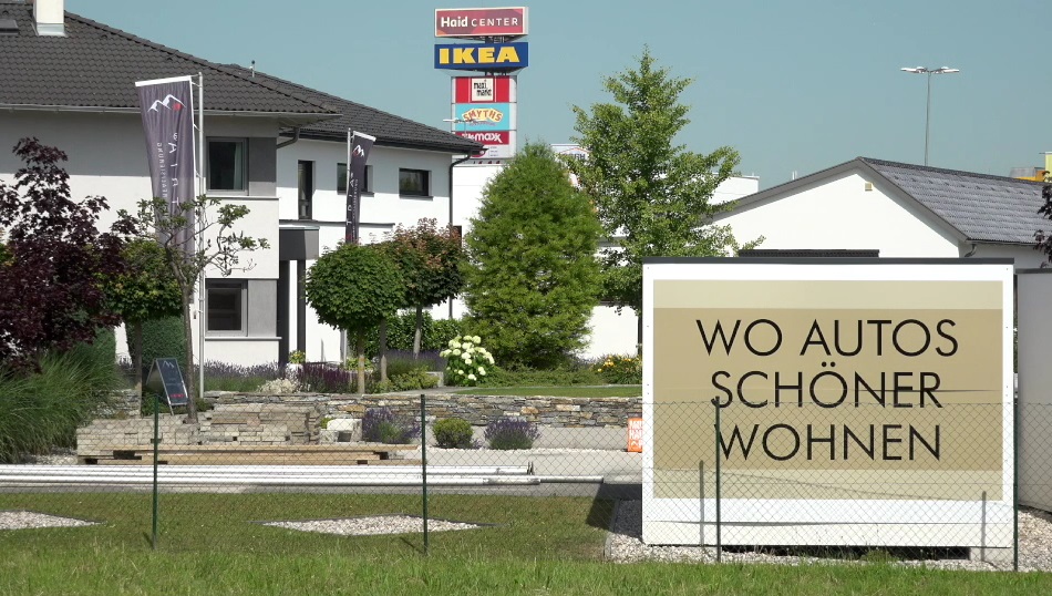 Schild mit der Schrift "Wo Autos schöner wohnen" vor einer Häuser landschaft